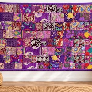Op de afbeelding: Een rechthoekige wandhanger met een patchworkontwerp van diverse stoffen vierkanten in tinten paars, magenta en goud. Het textiel toont ingewikkelde bloemen-, paisley- en geometrische patronen met geborduurde details en een paarse rand.