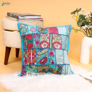 May include: A square throw pillow featuring a vibrant patchwork design with various embroidered floral and paisley patterns in shades of turquoise, pink, red, and gold. The pillow is finished with a bright turquoise border and intricate stitching.