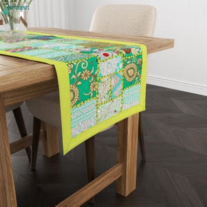Può includere: Un runner da tavola rettangolare caratterizzato da un bordo verde brillante e un design patchwork composto da vari quadrati di tessuto. I quadrati presentano intricati motivi floreali e geometrici nei toni del bianco, rosso e oro, con cuciture visibili.