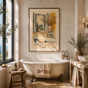 Puede incluir: Una lámina de arte enmarcada titulada BAIN DE LUMIERE que muestra una ilustración de un baño con una ventana abierta al mar, una bañera y un jarrón con flores. La obra utiliza tonos cálidos amarillos, naranjas y azules sobre un fondo crema.