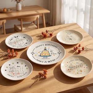 Puede incluir: Cinco platos redondos de cerámica con diversas ilustraciones. Uno muestra un calendario de septiembre, otro un boceto de boda, uno un pastel de cumpleaños con 'Happy Birthday Make a Wish', uno un calendario de diciembre con un gato y otro un pastel.