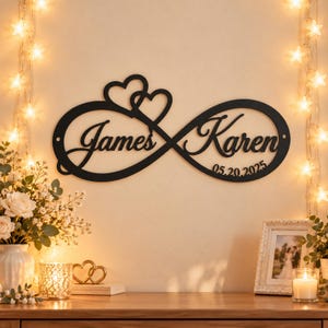 May include: A black metal wall sign shaped like an infinity symbol with two hearts at the top. The sign features the names James and Karen in cursive script, along with the date 05.20.2025 integrated into the design.