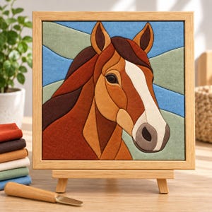 Op de afbeelding: Een vierkant wandkunstwerk van stof met een gestileerde paardenkop in tinten bruin, beige en wit, tegen een geometrische blauwe en groene achtergrond. Het kunstwerk zit in een lichtgekleurde houten lijst en staat op een kleine ezel.
