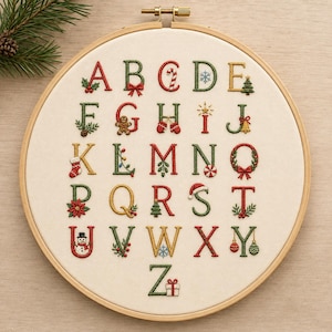May include: A circular wooden embroidery hoop containing a cream-colored fabric panel with a hand-stitched alphabet. Each letter is decorated with festive holiday motifs including holly, candy canes, snowflakes, ornaments, and a snowman.