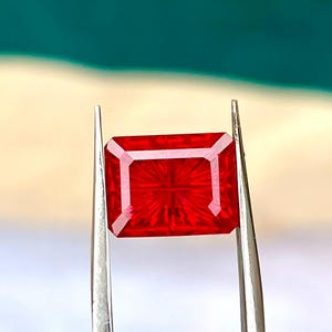 Peut inclure: Une pierre précieuse rouge vif de forme rectangulaire à taille à degrés, présentant des facettes géométriques précises créant un motif rayonnant depuis le centre. La pierre est tenue par des pinces en métal argenté, révélant sa couleur cramoisie.