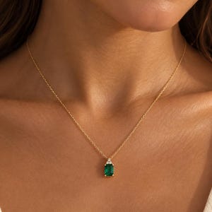 May include: A gold chain necklace featuring a rectangular emerald-cut green gemstone pendant. The pendant is accented with two small, clear, round-cut stones positioned at the top, creating a delicate and refined piece of jewelry.