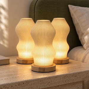 May include: Three table lamps with white, vertically ribbed, hourglass-shaped shades. Each lamp features a warm, glowing light source and sits on a circular, light-colored wooden base. The lamps are arranged in a cluster on a textured surface.
