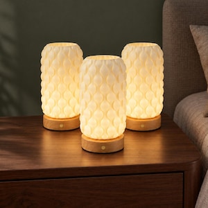 May include: Three cylindrical table lamps with off-white, textured, teardrop-patterned shades. Each lamp features a circular light-colored wooden base with a small, centered circular button on the front, emitting a warm, soft glow.