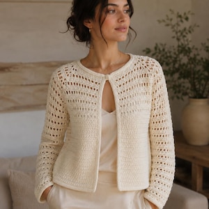 May include: A cream-colored, long-sleeved crochet cardigan featuring an open-knit pattern on the sleeves and upper bodice, with a solid knit lower section and a single button closure at the neckline, worn over a matching cream-colored camisole.