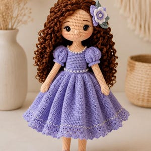 May include: A handcrafted crochet doll with long, curly brown hair and a light skin tone. The doll wears a lavender dress with puff sleeves, a pearl-accented neckline and waist, and a scalloped hem. A small purple flower accessory is in the hair.