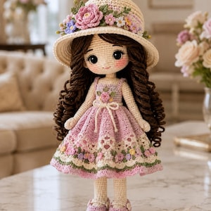 May include: A handmade crochet doll with long brown curly hair, wearing a cream-colored sun hat adorned with pink and purple flowers. The doll wears a light pink dress featuring a floral pattern, a white lace-like hem, and matching pink shoes.