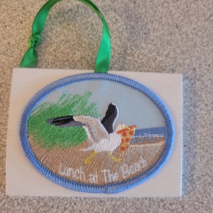 May include: Embroidered rectangular ornament with a green ribbon. The oval patch features a seagull carrying pizza on a beach with the text "Lunch at The Beach". The patch is bordered with blue stitching.