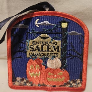 May include: Embroidered patch with a blue background and a sign that reads "Entering Salem Massachusetts". The sign is surrounded by vines and a street lamp. There are three jack-o'-lanterns in the foreground.