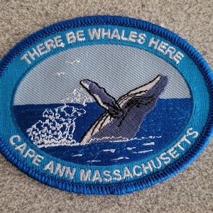 May include: Blue and white embroidered patch with a whale tail breaching the water. The text reads "There Be Whales Here Cape Ann Massachusetts."