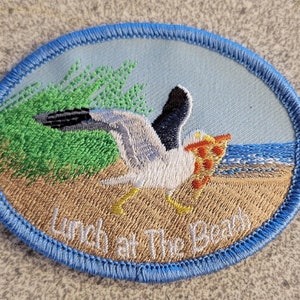 May include: Embroidered patch with a blue border, depicting a seagull holding a slice of pizza on a beach with green grass. The text "Lunch at the Beach" is embroidered below the seagull.