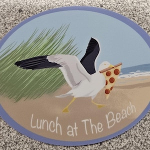 May include: Oval sticker featuring a cartoon seagull carrying a slice of pizza on a beach. The seagull is white and gray with black wingtips and yellow feet. The background shows a beach scene with text that reads "Lunch at The Beach."