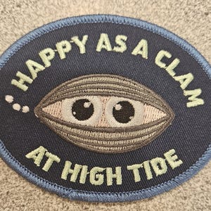 May include: Navy blue embroidered patch with a light blue border. The patch features a cartoon clam with large eyes and the text "HAPPY AS A CLAM AT HIGH TIDE" in light green lettering. The design is oval-shaped.