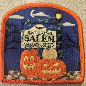 May include: An embroidered patch with a blue background and orange border. The patch features a sign that reads "Entering Salem Massachusetts" with a bat flying above it. There are three pumpkins in the foreground, two orange and one beige, with jack-o-lantern faces.