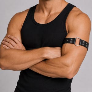 Può includere: Un bracciale da braccio in pelle nera caratterizzato da un anello centrale in metallo color argento e due file di borchie metalliche color argento. La fascia è indossata intorno alla parte superiore del braccio sopra una canotta nera.