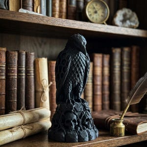 Pode incluir: Uma estatueta de corvo preto fosco pousada sobre uma base decorada com vários crânios humanos e entalhes intrincados em forma de videira. O pássaro apresenta texturas de penas detalhadas e um bico afiado, como um objeto escultural escuro.