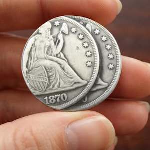 Może przedstawiać: Dwie srebrzyste monety przedstawiające siedzącą postać trzymającą laskę i tarczę z napisem LIBERTY. Monety są datowane na 1870 rok i ozdobione rzędem gwiazd wzdłuż górnej krawędzi, posiadają metaliczne wykończenie.