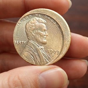 Może przedstawiać: Miedziana moneta jednocentowa Lincoln z 1909 roku z literą S, przedstawiająca profil Abrahama Lincolna zwrócony w prawo. Napis IN GOD WE TRUST jest wygięty wzdłuż górnej krawędzi, słowo LIBERTY znajduje się po lewej, a data 1909 ze znakiem S po prawej.
