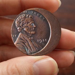Può includere: Una moneta color rame con un ritratto di profilo di Abraham Lincoln rivolto verso destra. Il testo WE TRUST è visibile in alto e l'anno 1914 con un segno di zecca S è stampato a destra del ritratto. La moneta ha una forma circolare.