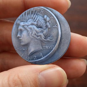 Könnte beinhalten: Ein kreisförmiges, silberfarbenes Objekt mit einem Relief des Liberty-Kopfes, das an einen Peace-Dollar von 1924 erinnert. Die Oberfläche zeigt das Wort LIBERTY oben, den Schriftzug IN GOD WE TRUST unten sowie die Jahreszahl 1924.