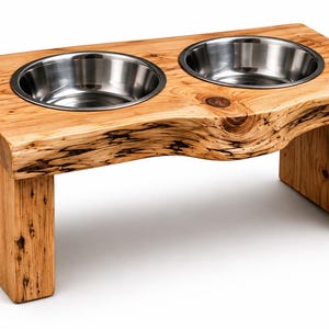 May include: A raised pet feeder stand crafted from natural light brown wood with a live edge front, featuring two circular stainless steel bowls set into the top surface. The wood grain is visible with dark knots and textured patterns throughout.