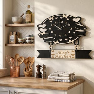 Puede incluir: Un reloj de pared negro con números blancos, que presenta recortes decorativos de mariposas, corazones y tazas de té apiladas. Un letrero de madera rectangular cuelga debajo del reloj con el texto Alice's Kitchen y detalles de corazones.