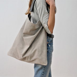 Könnte beinhalten: Eine große, hellgraue Tragetasche mit Schulterriemen. Die Tasche ist aus einem weichen, Wildleder-ähnlichen Material gefertigt und hat ein schlichtes, minimalistisches Design. Der Riemen ist dunkelgrau und mit goldfarbener Hardware befestigt.