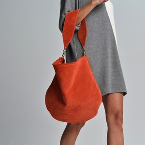 May include: An orange suede shoulder bag with a wide strap and gold-colored hardware. The bag has a rounded shape and a soft, textured appearance. The strap is also orange and appears to be suede.