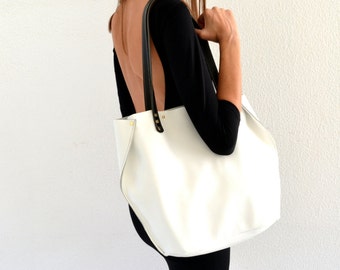 May include: A white leather tote bag with black leather handles and gold accents. The bag is being held by a person wearing a black dress and sunglasses.