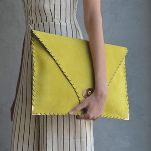 Può includere: Una pochette giallo brillante con chiusura a patta e chiusura dorata. I bordi sono cuciti con filo blu scuro. La pochette è tenuta davanti a una persona che indossa un vestito a righe.