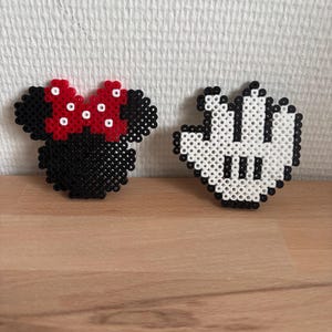 Peut inclure: Deux figurines en pixel art réalisées avec des perles en plastique. L'une représente une silhouette de tête de souris noire avec un nœud rouge à pois blancs. L'autre est un gant de dessin animé blanc avec des contours et trois lignes noires.