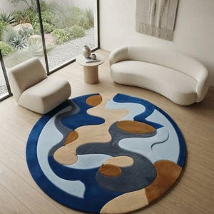 May include: A large circular area rug featuring an abstract pattern of organic, flowing shapes in shades of deep blue, light blue, tan, and dark gray. The rug has a textured, multi-level pile that creates depth and dimension across its surface.