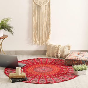 Könnte beinhalten: Ein runder roter Wandteppich mit einem detaillierten Mandala-Muster in Orange und Pink, das komplexe Blumen- und Paisley-Motive aufweist. Der Stoff liegt auf dem Boden und dient als Unterlage für einen Laptop, eine Tasse und zwei Bücher.