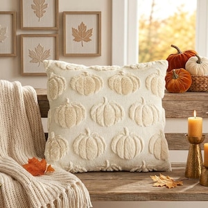 May include: A square cream-colored throw pillow featuring a raised, textured pumpkin pattern across the front. The pillow is displayed alongside a cream knit throw blanket, several small decorative pumpkins in a basket, and four gold-toned candle holders.