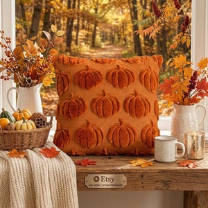 May include: A square throw pillow in a burnt orange color featuring a repeating pattern of raised, fuzzy pumpkin shapes. The pillow is displayed on a wooden surface alongside a wicker basket of gourds, a white mug, and a candle.