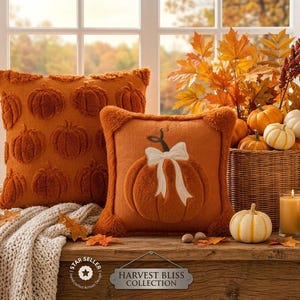 May include: Two square orange throw pillows, one featuring a repeating tufted pumpkin pattern and the other a single large tufted pumpkin with a white ribbon bow. A wicker basket holds small pumpkins and autumn leaves near two lit pillar candles.