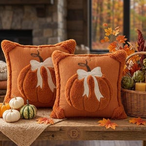 May include: Two square orange throw pillows featuring a textured, raised pumpkin design with a white embroidered bow. The pillows have a fuzzy, tufted border and are displayed alongside small decorative pumpkins and a woven basket of autumn decor.