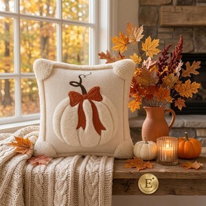 May include: A square cream-colored knit throw pillow featuring a plush, raised white pumpkin applique with a dark brown stem and a burnt orange bow. The pillow has soft, fuzzy corner accents and is displayed with autumn decor and a candle.