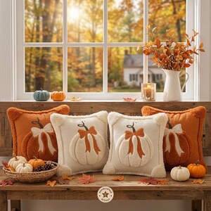 May include: Four square throw pillows featuring raised pumpkin designs with bows. Two pillows are orange with white pumpkin appliques, and two are cream with orange pumpkin appliques. A woven basket holds small pumpkins, and a white pitcher holds branches.