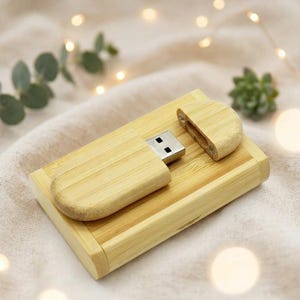May include: A light-coloured bamboo USB flash drive with a matching rectangular bamboo storage box. The drive features a removable cap and a silver metal connector, resting on the lid of the box which has a smooth, natural wood grain finish.
