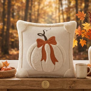 May include: A square cream-colored knit throw pillow featuring a plush, raised white pumpkin design in the center. The pumpkin is accented with a dark brown stem and a prominent, textured orange bow tied around the top of the pumpkin shape.