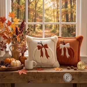 May include: Two square plush pillows featuring raised pumpkin designs with bows. One pillow is cream with a rust-colored bow, and the other is rust-colored with a cream bow. They sit on a wooden surface with a ceramic mug and small pumpkins.