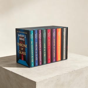May include: A complete book collection box set titled Throne of Glass by Sarah J. Maas. The set includes eight books with colorful spines in shades of teal, blue, purple, green, red, orange, and black, featuring gold text and a dark blue box.