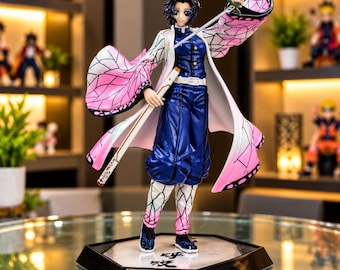 Puede incluir: Una figura de anime de Shinobu Kocho sobre una base hexagonal con texto en japonés. El personaje viste un uniforme azul oscuro, un haori blanco con patrón de alas de mariposa rosa y negro, y sostiene una espada fina. Pelo negro.