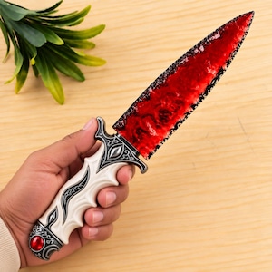 May include: A decorative dagger featuring a translucent red blade with a textured, dark-edged finish. The handle is cream-coloured with intricate silver-toned metal filigree, a central eye-shaped accent, and a round red gemstone at the pommel base.