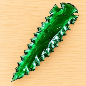 Könnte beinhalten: Eine leuchtend smaragdgrüne Pfeilspitze aus Glas mit einer scharfen, gezackten Kante auf beiden Seiten und einer spitzen Spitze. Die Oberfläche weist ein glattes, glänzendes Finish mit subtilen internen Wirbeln und Transparenz auf.